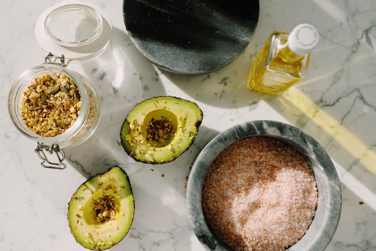 about-us Top view of tasty halved avocado sprinkled with various seeds placed near pink salt with seed mixture in glass pot and other dressing ingredients arranged on white marble table