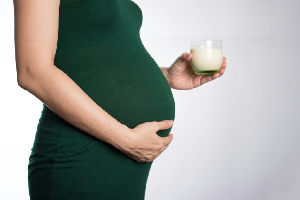 Close-up of a pregnant woman in a green dress holding a glass of milk in a studio setting.